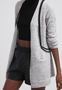 Grå cardigan med avslappnad passform, med svarta kantband, långa ärmar och framfickor, lager på lager över en svart skjorta och lädershorts.