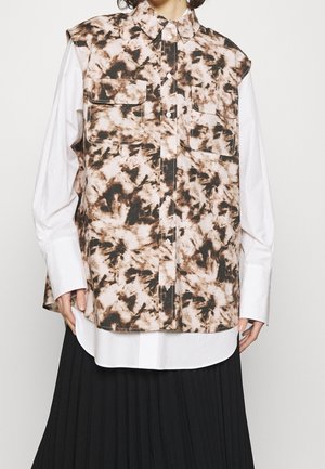 Gilet sans manches marron et noir à motif tie-dye avec poches avant, porté sur une chemise blanche à manches longues boutonnée et une jupe plissée noire.