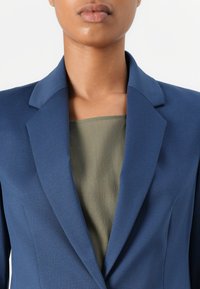 Tummansininen bleiseri, joka on muotoiltu istuvaksi, notkatuilla kauluksilla ja sileällä pinnalla, kerroksittain khaki-vihreän yläosan päällä. Lähikuvassa ylävartalo.