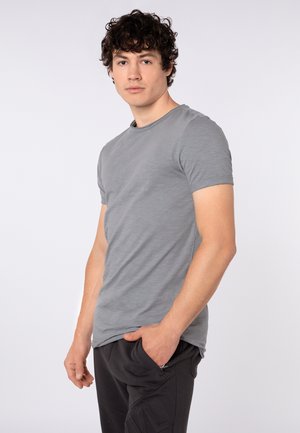 Junger Mann mit lockigem Haar, der ein graues kurzärmeliges T-Shirt und schwarze Hose trägt, steht mit einer Hand in der Tasche vor einfachem Hintergrund.