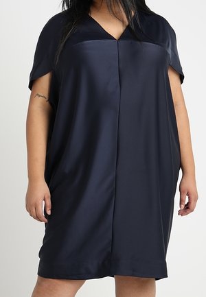 Frau trägt ein lockeres, kurzärmeliges, dunkelblaues Kleid mit V-Ausschnitt und einem vertikalen Mittelnaht vorne.