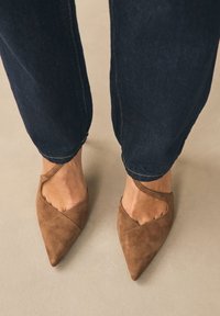 Rudos zomšiniai aukštakulniai batai su smailiomis galais ir asimetrišku pjūviu, su dirželiu aplink kulkšnį. Dėvimi su tamsiais džinsais.