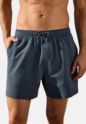 Man wearing dark blue textured swim shorts with drawstring waist and side pockets, standing with hands near pockets.