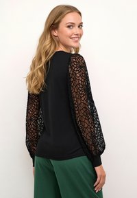 Black long-sleeve top featuring sheer lace sleeves with a floral pattern, relaxed fit, and ribbed cuffs, paired with green pants.