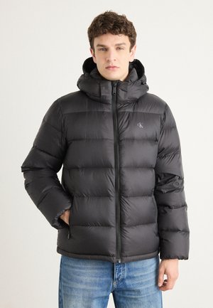 Black puffer jacket with a high collar and hood, featuring a zip front, quilted texture, and logo accent on the chest.
