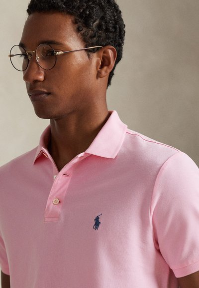 Ροζ πόλο μπλούζα με υφασμάτινη υφή, με μικρό μπλε λογότυπο στο αριστερό στήθος, τρία κουμπιά και κλασικό γιακά.