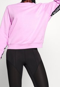 Sudadera rosa con cuello redondo y franjas de acento negras que presentan texto a lo largo de las mangas, combinada con leggins negros ajustados con paneles de malla.