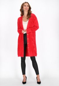 Helle rote, flauschige lange Strickjacke mit goldenen Knöpfen und verzierten Details an den Schultern, kombiniert mit schwarzen Lederleggings und hohen Schuhen.