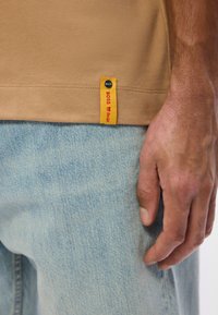 Μπεζ T-shirt με ευθεία άκρη και κίτρινη ετικέτα που εμφανίζει το "BOSS" σε κόκκινο χρώμα. Φορεμένο με ανοιχτό γαλάζιο τζιν, που δείχνει την υφή του υφάσματος.