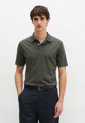 Junger Mann trägt ein dunkelgrünes Poloshirt und einen schwarzen Gürtel mit marineblauen Hosen, steht vor einem schlichten weißen Hintergrund.