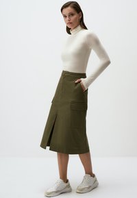 Olijfgroene knielange rok met een verticale split aan de voorkant, grote zijzakken en een aansluitende tailleband, gecombineerd met een crèmekleurige coltrui en sneakers.