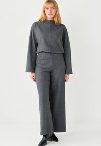 Blonde Frau, die ein dunkelgraues Langarmoberteil mit weitem Bein passende Hose und schwarze Stiefeletten mit Absatz trägt, steht vor weißem Hintergrund.