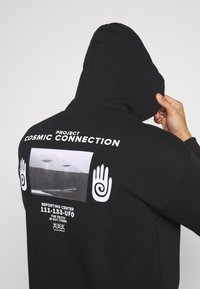 Felpa nera con cappuccio con stampe grafiche sulla schiena, tra cui "PROGETTO CONNESSIONE COSMICA," immagini di UFO e design di mani emblematiche.