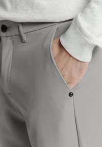 Grijze op maat gemaakte broeken met een gladde textuur, voorzien van een zijzak en een donkere knoopsluiting nabij de tailleband, gedragen met een lichte trui.