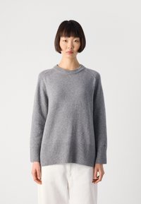 Frau mit kurzem dunklem Haar, die einen hellgrauen Langarmpullover und weiße Hosen trägt und vor einem schlichten weißen Hintergrund steht.