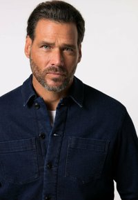 Tmavě modrá denimová košile s knoflíkovým zapínáním vpředu, dvěma kapsami na hrudi a límcem. Má strukturovanou tkaninu a černé knoflíky.