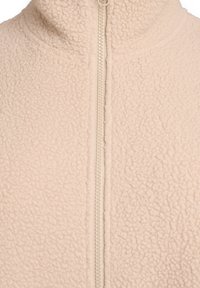 Beige fleecejack met een hoge kraag en een ritssluiting aan de voorkant, met een gestructureerd, zacht oppervlak en een minimalistisch ontwerp.