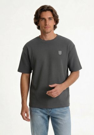Jeune homme aux cheveux mi-longs portant une chemise gris foncé à manches courtes avec un petit logo blanc, associée à un jean bleu clair.