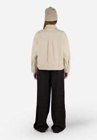 Giacca in fleece color crema con retro plissettato e polsini con bottoni, abbinata a pantaloni neri a gamba larga e un berretto di maglia beige.