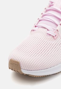 Zapatilla deportiva de color rosa claro con una parte superior de malla transpirable, un patrón texturizado y una suela de goma beige contrastante. Presenta cordones planos y un mínimo de marca.