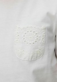 Camiseta blanca con un intrincado bolsillo blanco bordado que presenta patrones florales y circulares, creando un detalle de diseño texturizado.