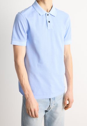 Mann trägt ein hellblaues kurzärmliges Polohemd mit schwarzen Knöpfen und hellblauen Jeans, steht vor einem einfarbigen Hintergrund.