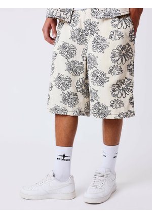 Persona che indossa pantaloncini color crema con motivi floreali neri, sneakers bianche Nike e calzini bianchi a metà polpaccio con loghi di aerei neri.