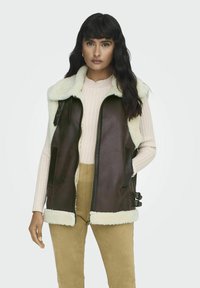 Bruine leren vest met een crèmekleurige nepbont kraag, zijzakken en verstelbare zijkolommen. Draagt over een lichtroze ribbeltrui.