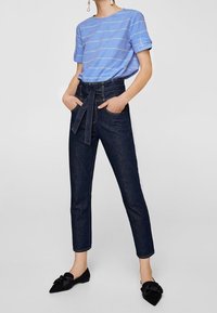 T-shirt rayé bleu, coupe décontractée, manches retroussées. Jean taille haute foncé avec un lien à l'avant. Ballerines noires avec un détail noeud.