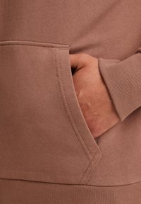 Sweatshirt rose poudré avec des poignets et un ourlet côtelés, présentant une grande poche avant avec un détail de couture en diagonale. Tissu doux et texturé.