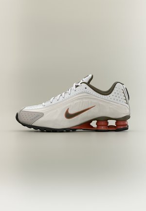 Weiß-grauer Nike Laufschuh mit orangefarbener federartiger Sohlenstütze und braunem Nike-Swoosh auf schlichtem neutralem Hintergrund.