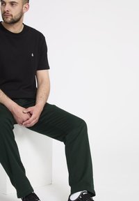 Zwarte katoenen t-shirt met een klein wit logo, gecombineerd met groene katoenen broek en zwarte sneakers, zittend op een witte kubus.
