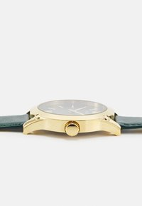 Gullur klokke med en mørk skjerm, grønn lærrem, rund form og teksturert kronedetaljering. Minimalistisk design med klare timemarkører.