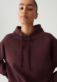 Donna con espressione neutra che indossa un hoodie bordeaux con lacci, in piedi di fronte a uno sfondo chiaro e semplice.