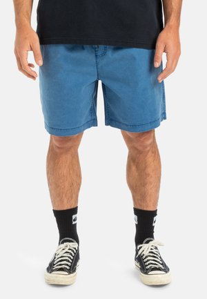 Blauwe denimshorts met zijknzippen, een relaxte pasvorm en een rechte zoom. Gecombineerd met zwarte sokken en klassieke zwarte sneakers.