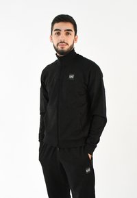 Chaqueta negra con cremallera fabricada en un material suave, que presenta un cuello alto y bolsillos laterales, combinada con joggers negros a juego y un pequeño parche con logo.