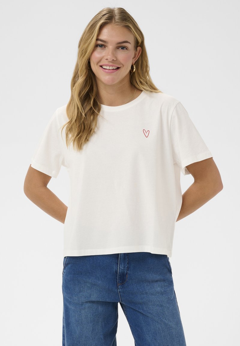 Usmievajúca sa žena s dlhými blond vlasmi, oblečená v bielom tričku s malým červeným srdcom na hrudi a modrých džínsoch, ruky za chrbtom.