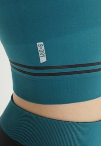 Primo piano di un top e un pantalone sportivo color teal che mostrano il logo B-DRY e strisce nere orizzontali su un tessuto testurizzato attorno alla vita.