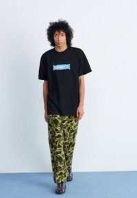 Camiseta negra con texto gráfico azul, combinada con pantalones de camuflaje en verde y marrón. El modelo lleva zapatillas negras y está de pie contra un fondo claro.