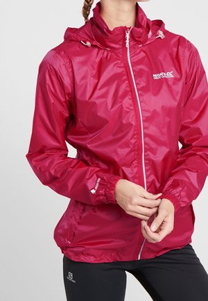 Femme portant une veste imperméable rose vif avec une capuche, ajustant le poignet, associée à des leggings noirs sur un fond uni.