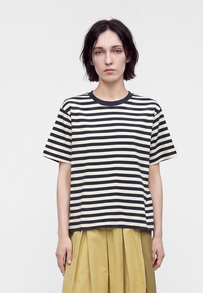 Jeune personne aux cheveux courts et foncés portant un t-shirt rayé noir et blanc et une jupe plissée beige, debout devant un fond uni.