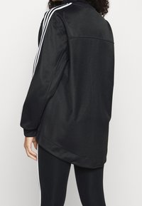 Svart atletisk jacka med vita ränder på axlarna, lätt material, avslappnad passform och rundad nederkant.