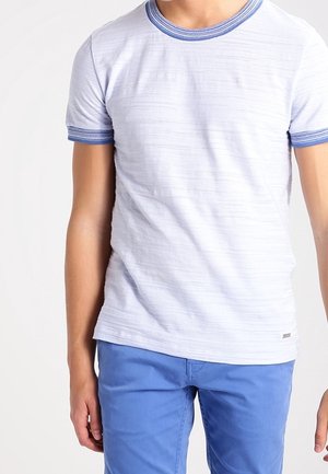 Man wearing a white short-sleeve t-shirt with blue trim on collar and sleeves, paired with blue pants, standing against a white background.