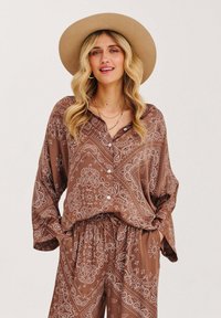 Hnědý vzorovaný outfit s košilí na knoflíky a kalhotami s elastickým pasem, vyrobený z měkké látky. Doplněno širokým béžovým kloboukem.