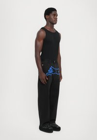 Sort tanktop parret med sorte jeans med et lyst blåt grafisk design. Modellen bærer mørke, broderede slip-on sko.