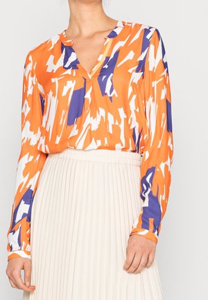 Blusa a maniche lunghe in un motivo astratto arancione, bianco e blu, con scollatura a V e tessuto morbido. Abbinata a una gonna plissettata bianca.