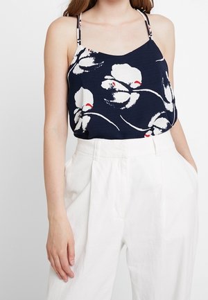 Woman wearing a navy blue floral spaghetti strap top tucked into high-waisted white wide-leg pants with hands in pockets.