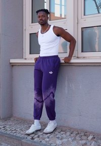 Calças roxas em tie-dye com punhos elásticos e um logotipo da adidas, combinadas com uma camisola branca e sapatilhas brancas. Tecido suave, ajuste casual.