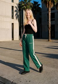 Jeune femme en débardeur noir et pantalon de survêtement vert avec des bandes blanches sur les côtés, souriant à l'extérieur près de palmiers et de bâtiments modernes.