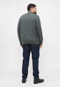 Pull en maille vert foncé avec une coupe décontractée, poignets et ourlet côtelés, associé à un jean en denim bleu foncé et des chaussures noires.